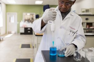 Scientist in a lab coat and gloves using a dropper to transfer blue liquid into a beaker in a laboratory setting.