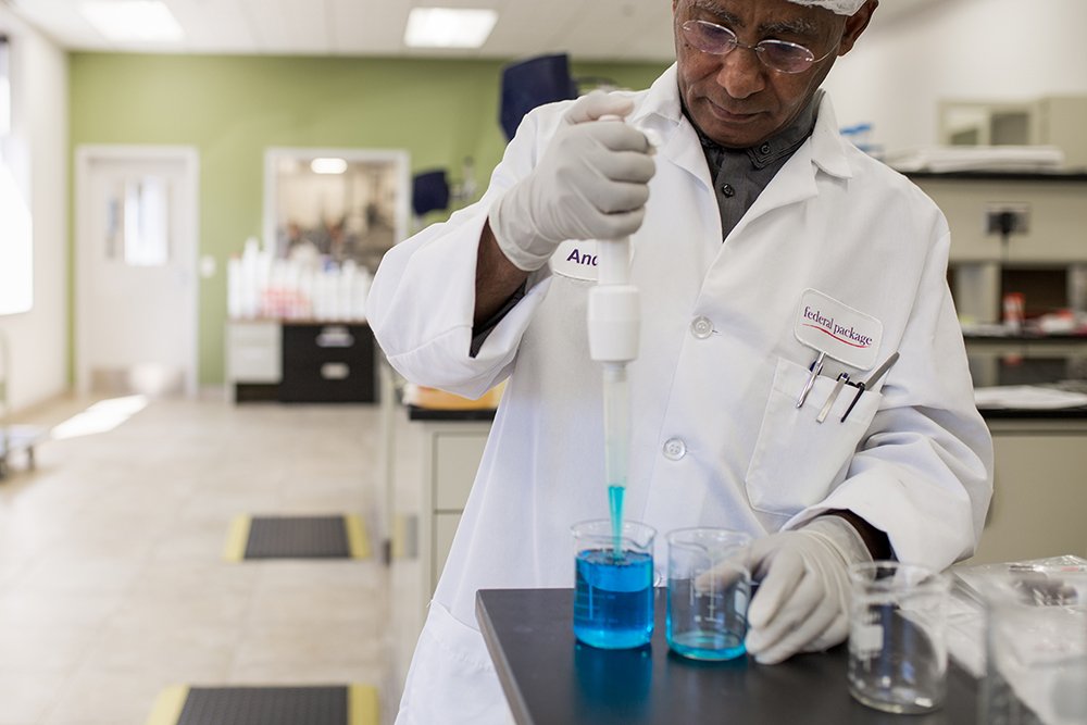 Scientist in a lab coat and gloves using a dropper to transfer blue liquid into a beaker in a laboratory setting.