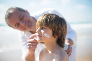 Father applying a custom sunscreen formulation lotion to his young son's cheek on a sunny beach.