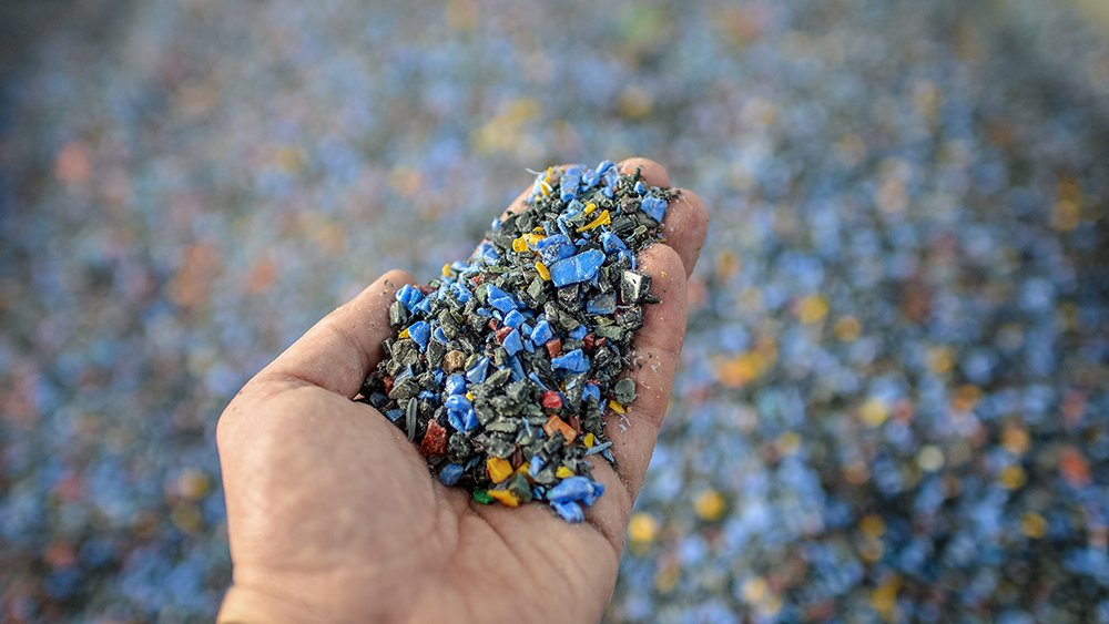 Close-up of a hand holding small multicolored pieces of recycled plastic, with a background covered in similar plastic fragments.