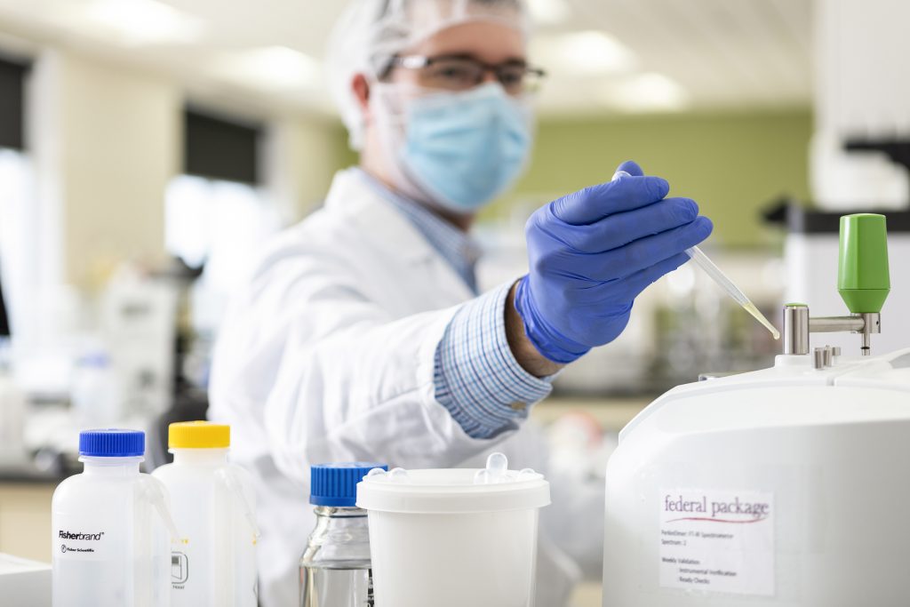 Lab technician wearing a face mask, hairnet, and gloves working with lab equipment in a laboratory, handling a pipette with various bottles and containers in the foreground.