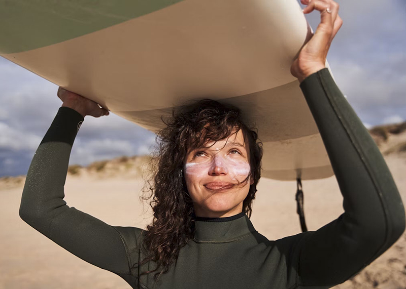 Woman holding a surfboard above her head with sun lotion streaks on her cheeks.