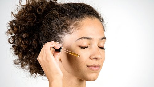 Close up of a woman's face with her eyes closed holding a dropper of serum. near her cheek bone.