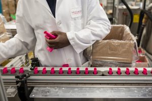 Worker in a Federal Package lab coat loading pink deodorant containers on an assembly line.