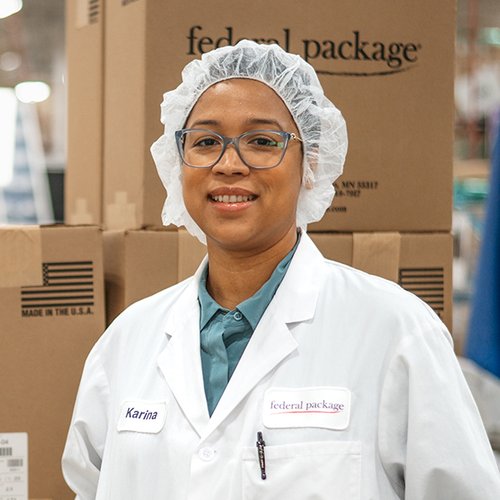 Portrait of a smiling woman wearing a lab coat and hair net standing in front of shipping boxes labeled federal package.