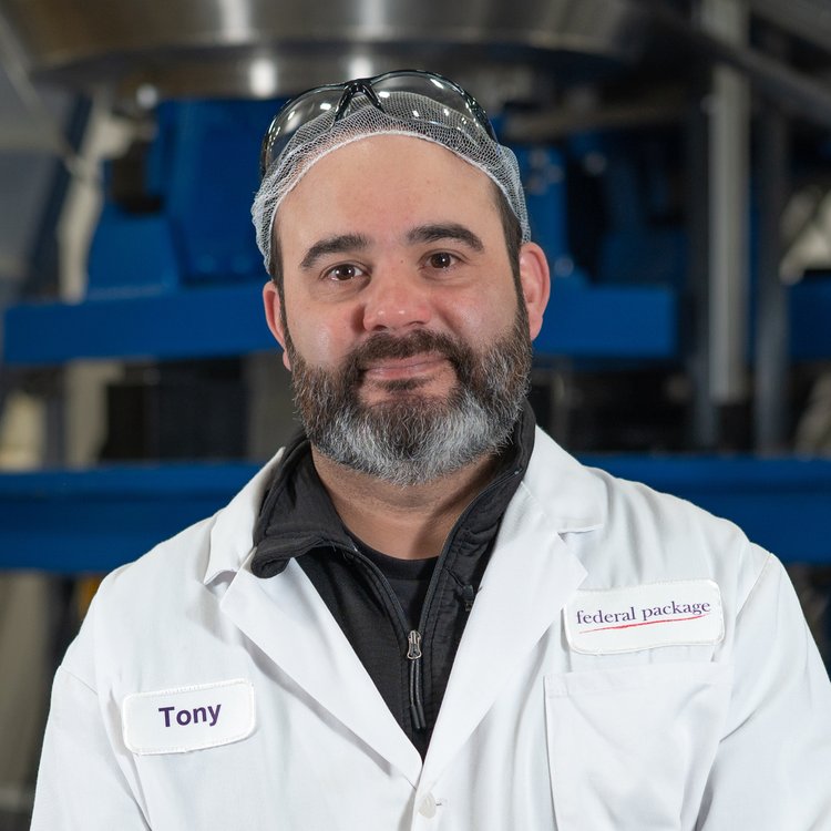 Close up of a man wearing a lab coat in a blurred industrial setting.