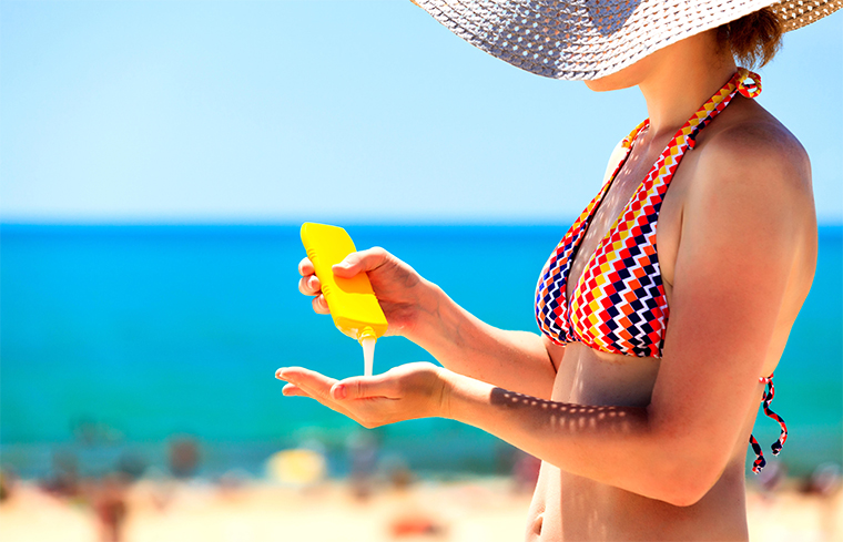 Woman wearing a sunhat and bathing suit pouring sunscreen into her hand with a blurred beach in the background.