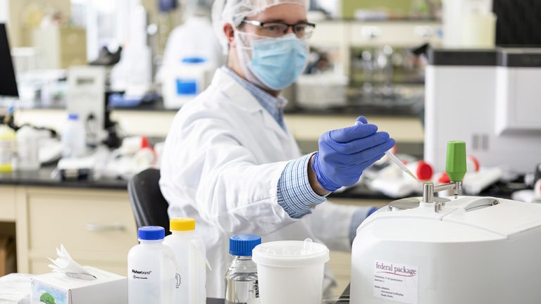 Lab technician wearing a face mask, hairnet, and gloves working with lab equipment in a laboratory, handling a pipette with various bottles and containers in the foreground.