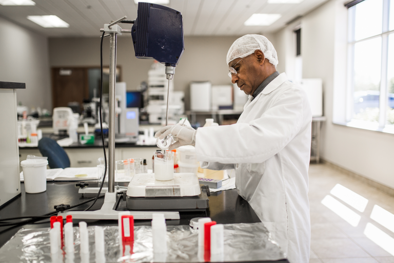 FP Lab scientist in a lab coat, hairnet, and safety glasses working with lab equipment to mix a white substance in a beaker inside a laboratory setting.