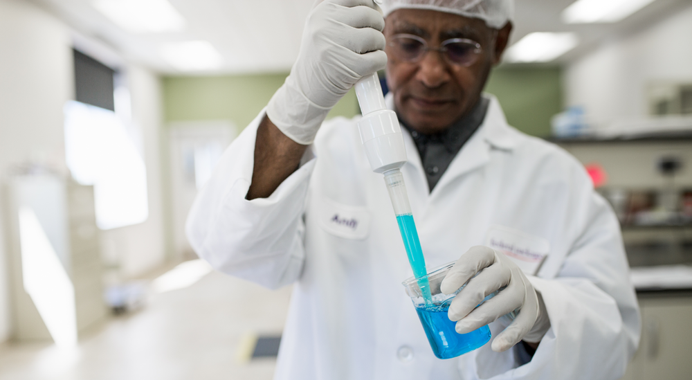 Scientist in a lab coat and gloves using a dropper to transfer blue liquid into a beaker in FP Lab's laboratory setting.