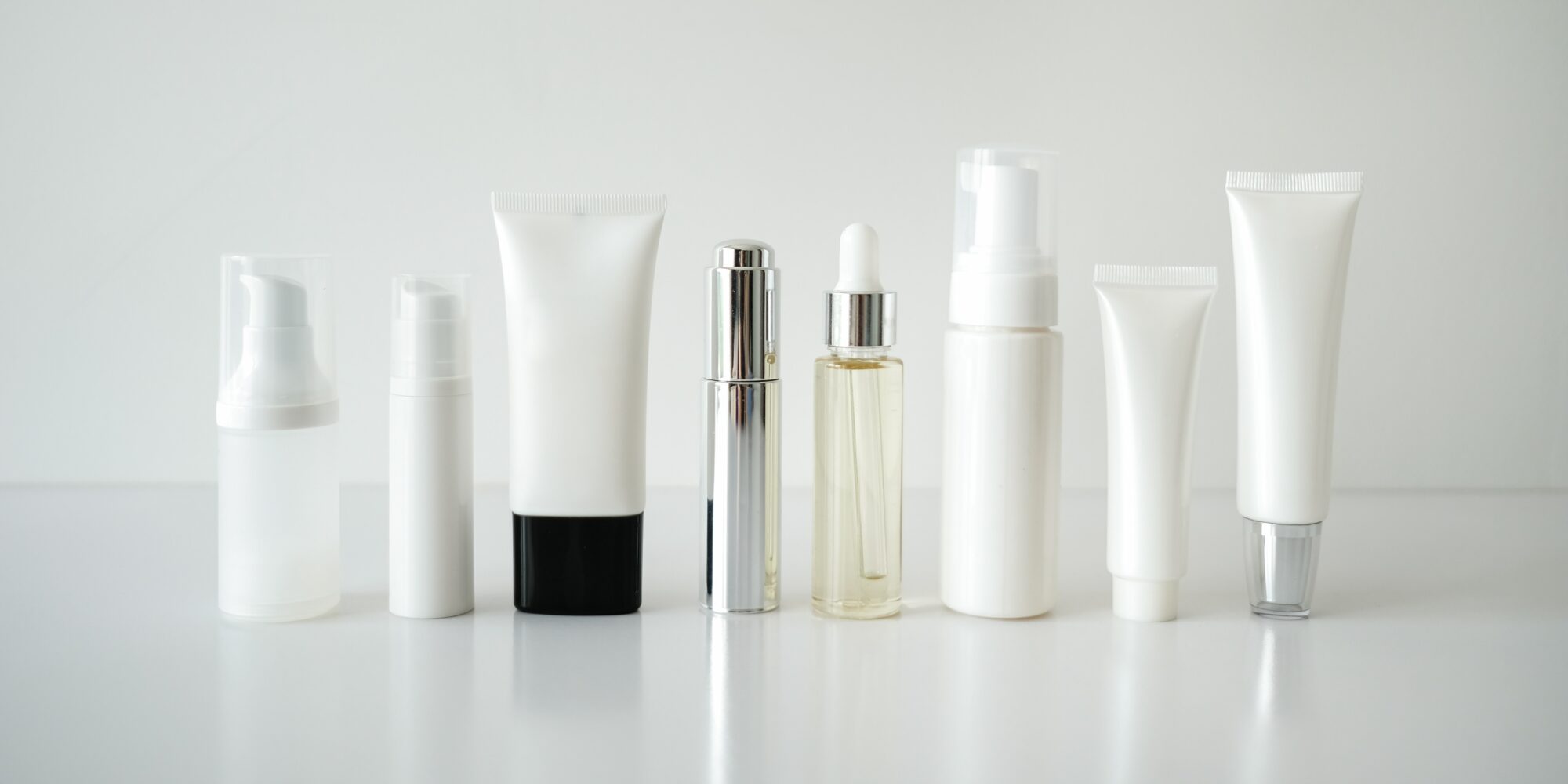 Assorted white and clear cosmetic containers, including bottles and tubes, displayed in a row on a white surface, representing skincare or cosmetic product packaging.