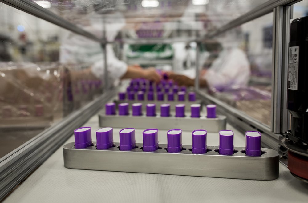 Close-up of a production line with several filled purple containers.Two out of focus people are working the assembly line in the background