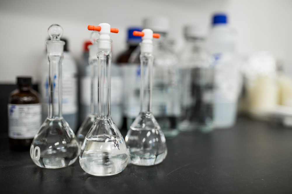 Clear laboratory flasks filled with liquid in a research and development lab, with various chemical bottles in the background.